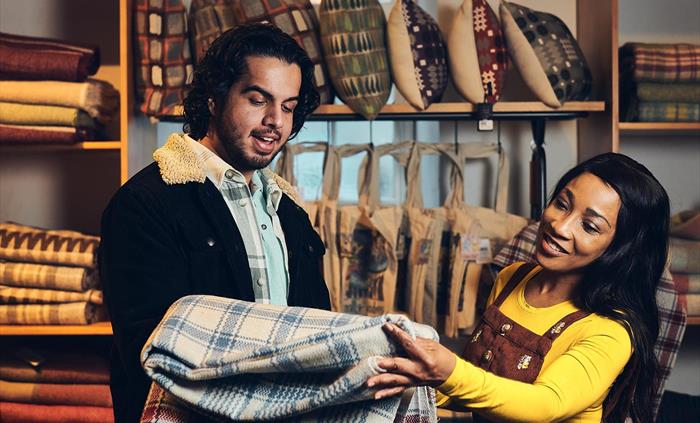 A couple stand in the museum shop, surrounded by blankets, cushions and bags. The man is holding up two woollen blankets as they decide which one to b
